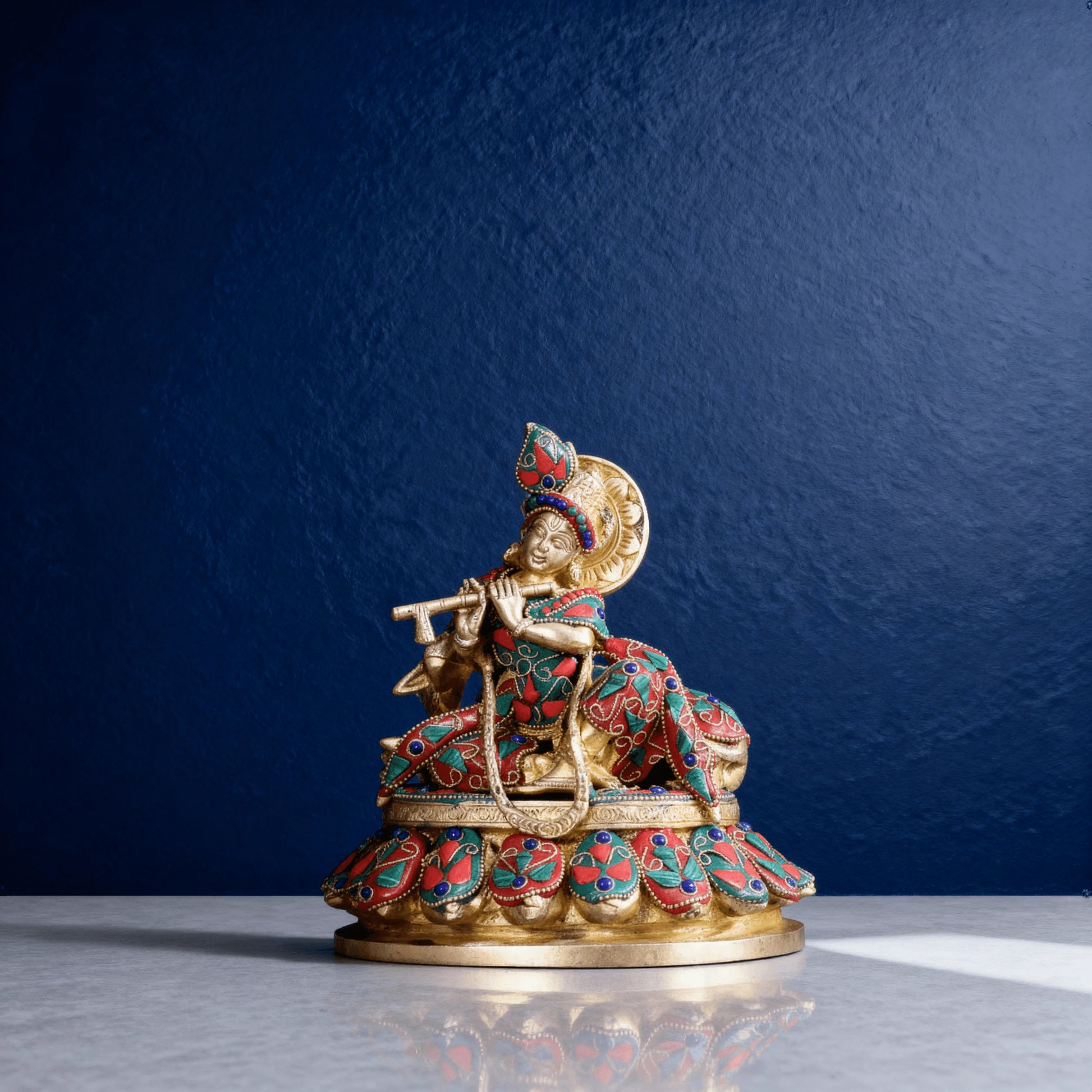 Handcrafted brass Krishna idol with meenakari detailing, seated on a decorated base, playing flute, side view.