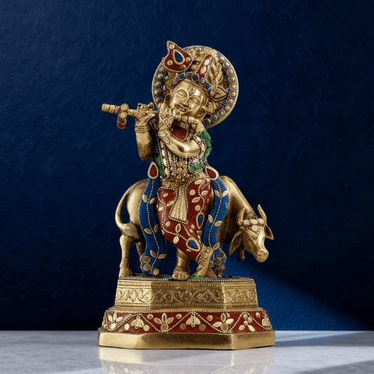Handcrafted brass Krishna with cow idol featuring red, blue and green stonework on a decorative base against dark blue background