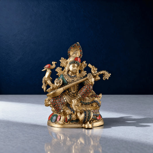 brass Saraswati idol with colorful stonework, photographed on a studio surface with a dark blue background and soft natural lighting