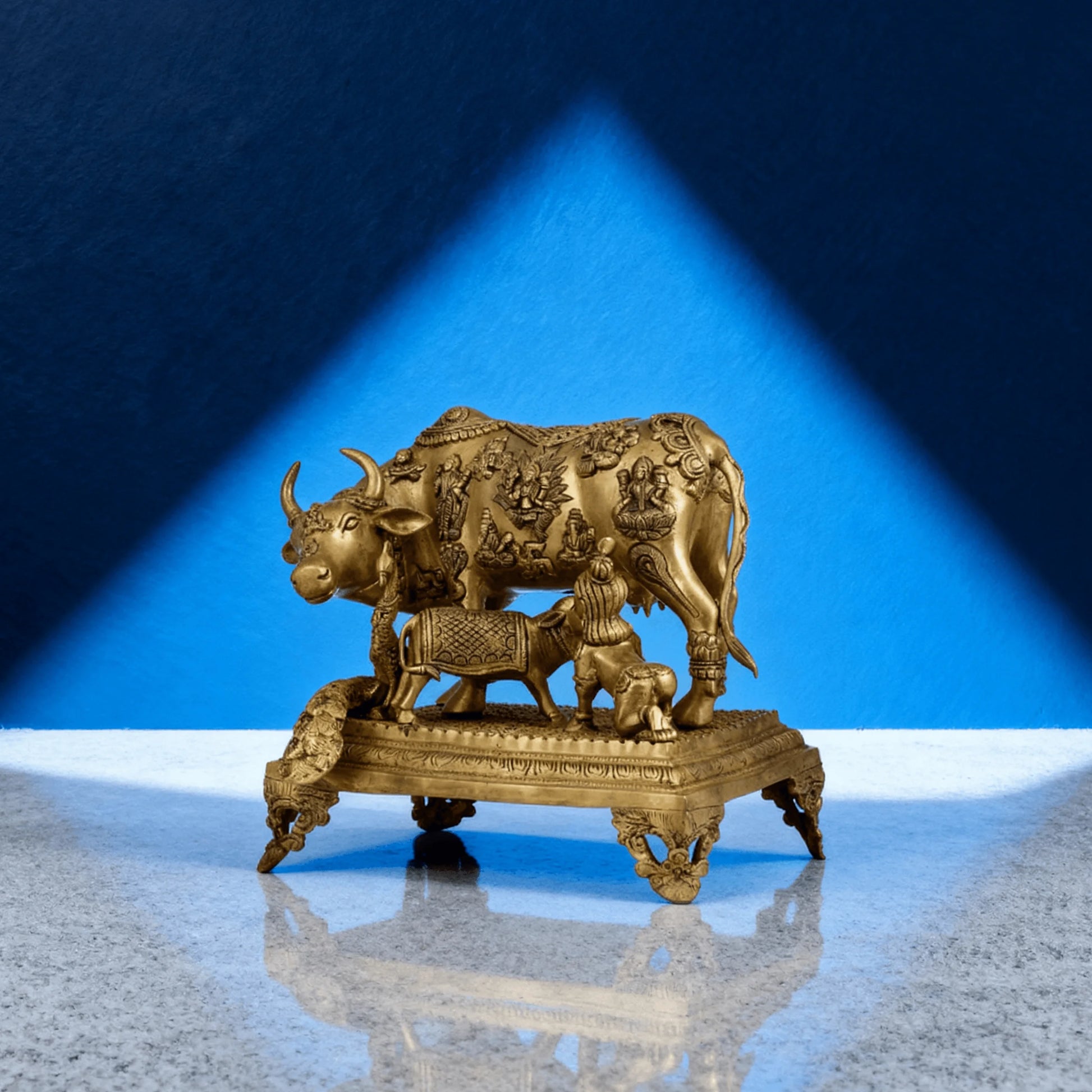Handcrafted pure brass Kamadhenu cow idol with calf, elevated on ornate base, shown against deep blue background