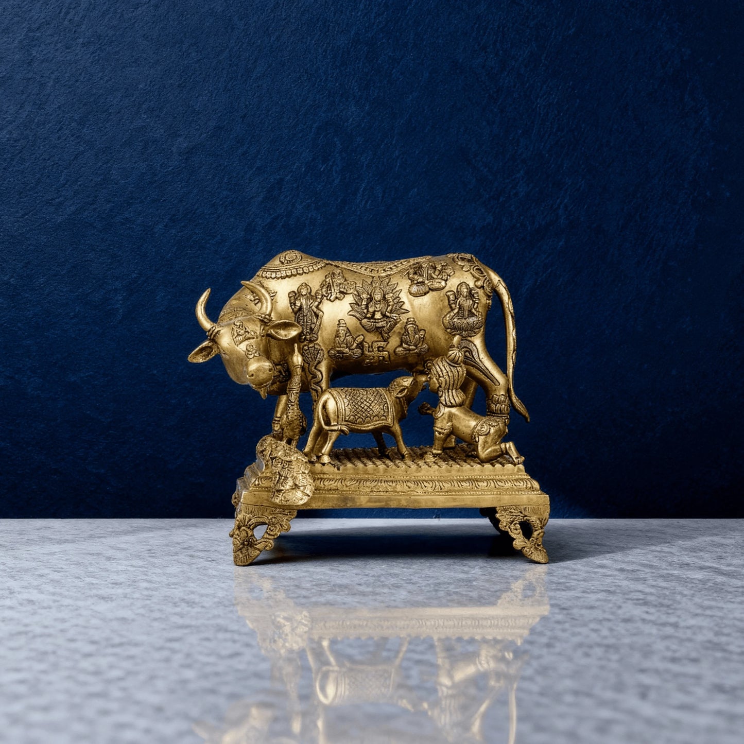 Brass Kamadhenu cow and calf idol in handcrafted detail, front perspective against dark blue wall