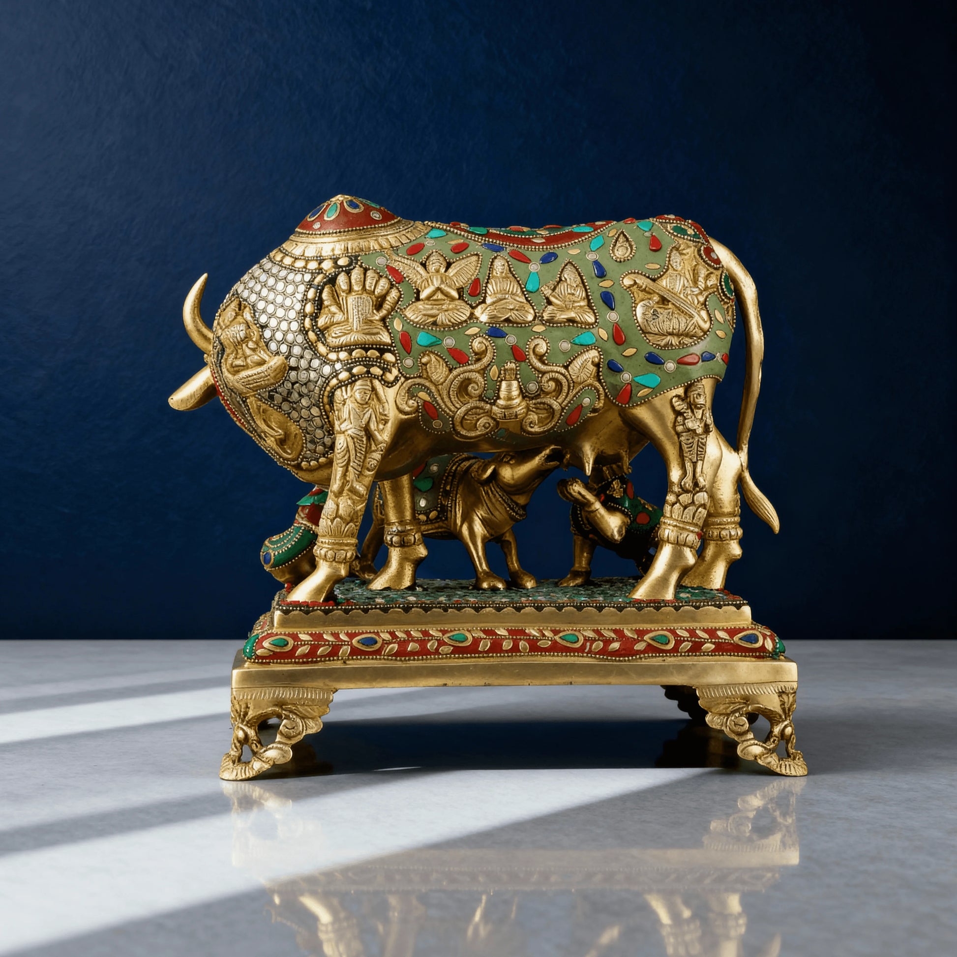 Rear view of detailed brass Kamadhenu idol with green and red stone inlay, intricate Indian motifs on a raised base