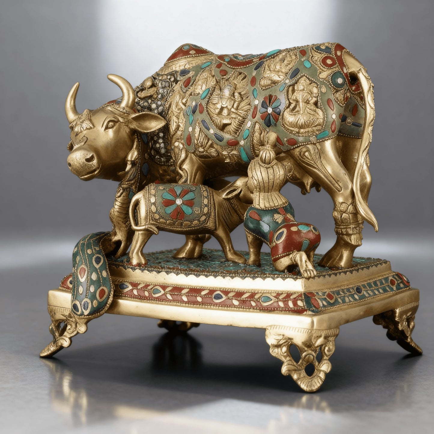 Close-up of brass Kamadhenu statue with calf and child, adorned with turquoise and red stones on ornate base