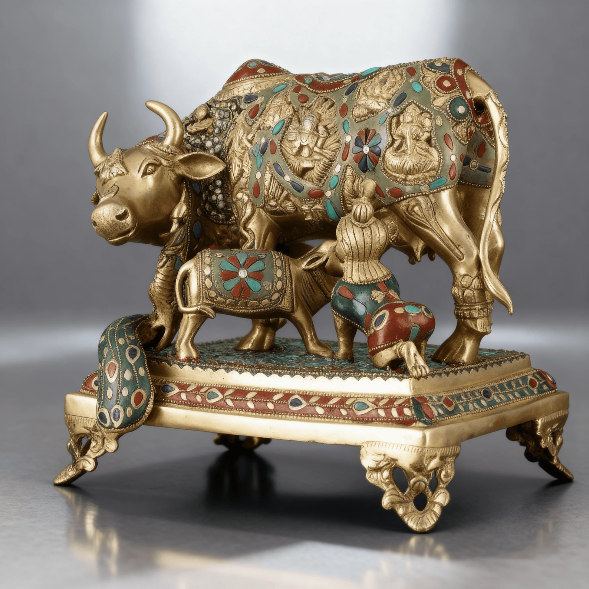 Close-up of brass Kamadhenu statue with calf and child, adorned with turquoise and red stones on ornate base
